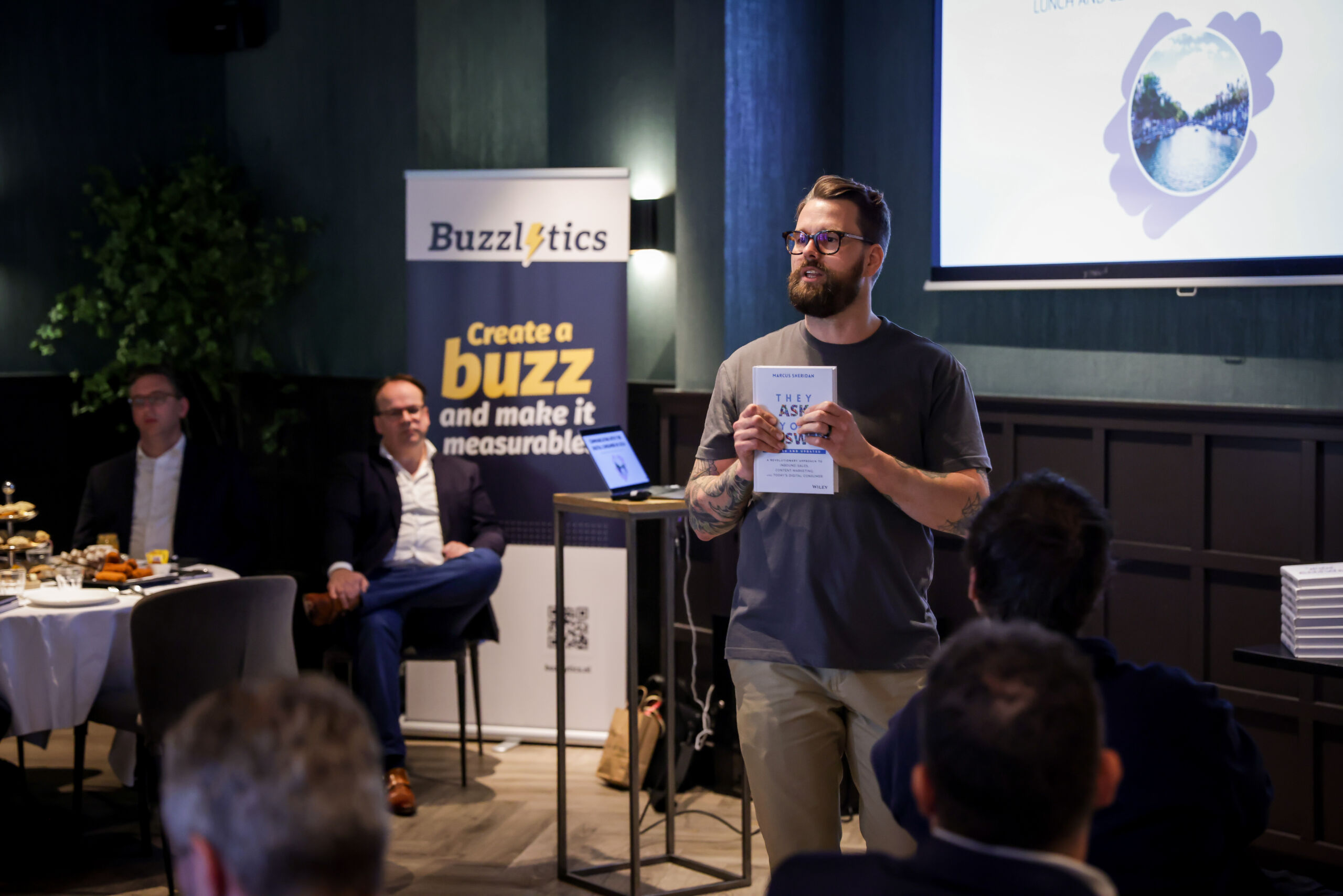 A man with a beard, glasses and a casual gray T-shirt stands speaking to an audience while holding a book. In the background is a banner reading "Buzzlytics" and the slogan "Create a buzz and make it measurable." Several people sit and listen intently, with some snacks visible on a table off to the side. The setting is a business meeting.