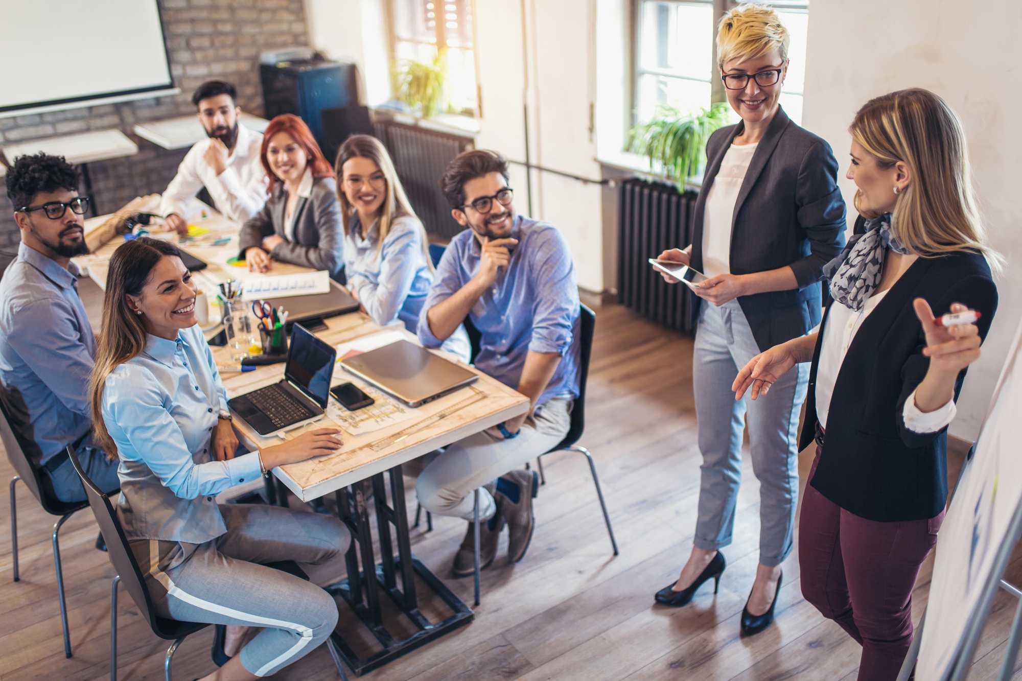 A diverse group of professionals in an office setting during a meeting. Seven people sit at a table with laptops and notebooks, while two women stand and lead the discussion. The atmosphere looks positive and engaged, with people smiling and listening intently.