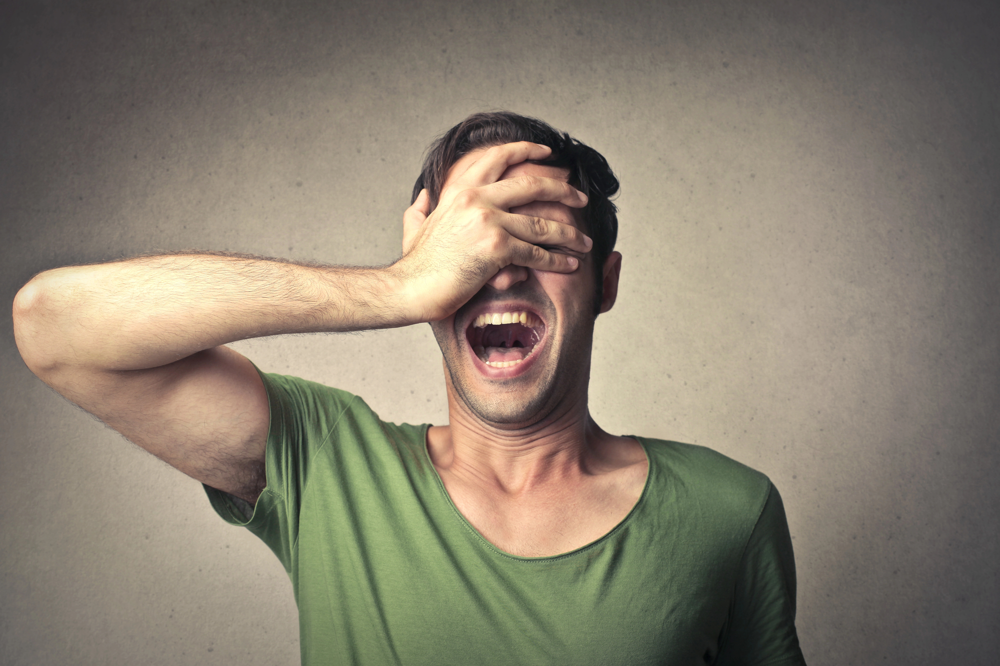 Man in green T-shirt covers his eyes with his hand against a gray background.