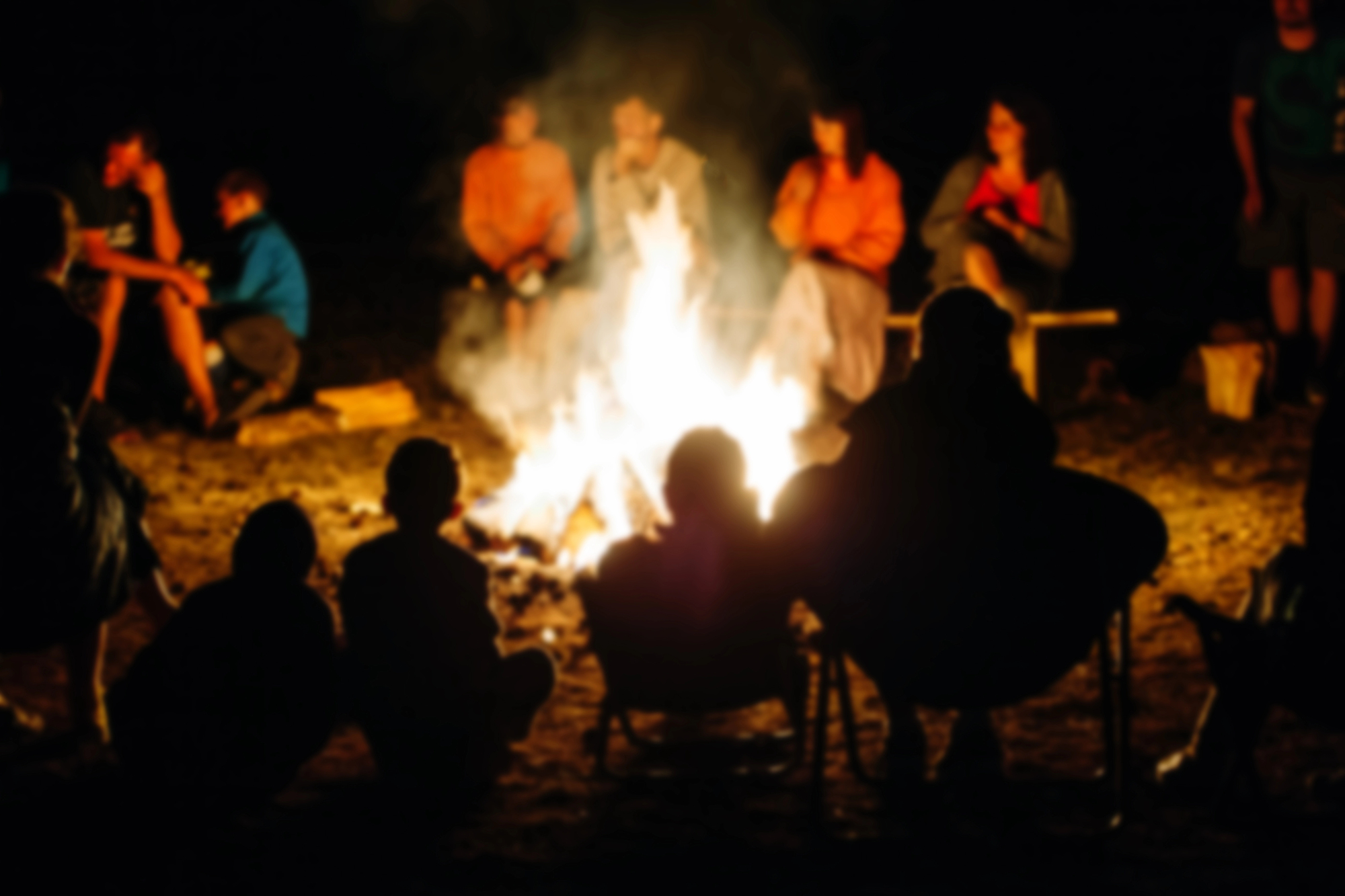 A group of people sit in a circle around a large campfire at night. The silhouettes of some people are visible in the foreground, while others are dressed in bright colors across the fire. The fire illuminates the surroundings with a warm glow.