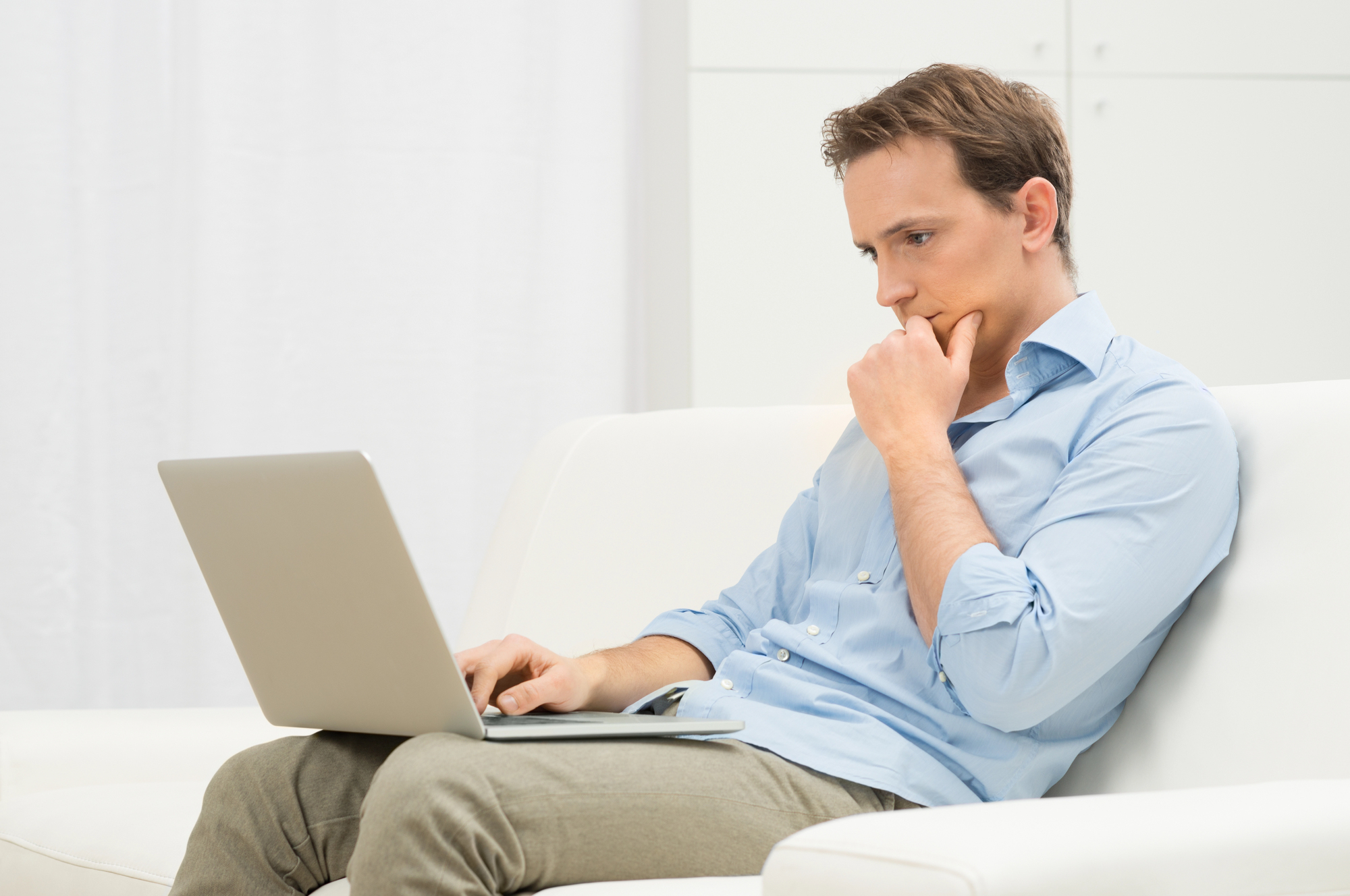 Man in light blue shirt sits on a white sofa with a laptop on his lap, hand by his chin, looking with a pensive expression as he contemplates a decision.