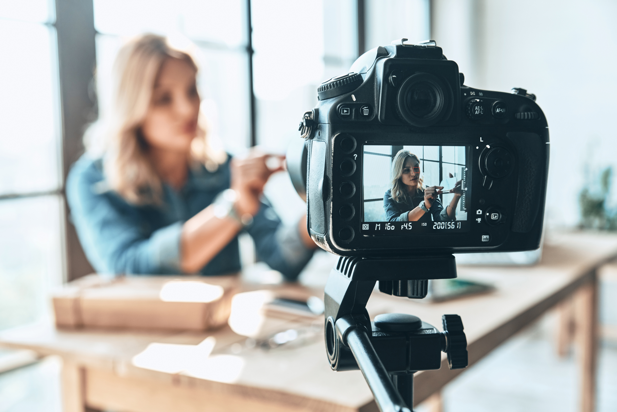 A blonde woman at a table, being filmed by a camera. She is wearing a denim blouse. The focus is on the camera display, which shows a bright image of the woman, while the background is out of focus. The room is well lit by large windows in the background.