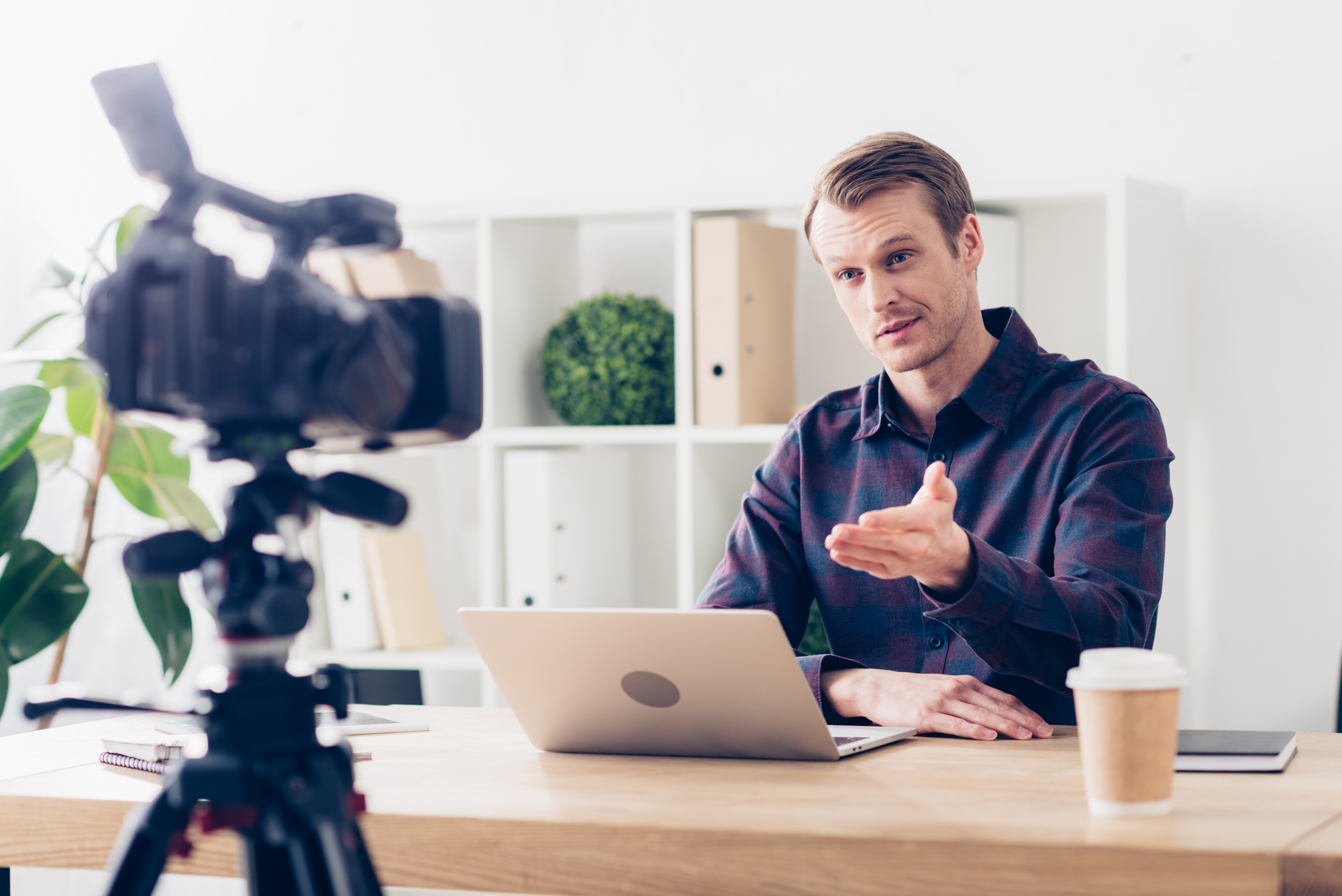 A man sits at a desk and talks in front of a camera. He is wearing a plaid shirt and has a laptop in front of him. Also on the desk are a paper cup with a lid and a notebook. Behind him, a number of shelves with books and plants can be seen.