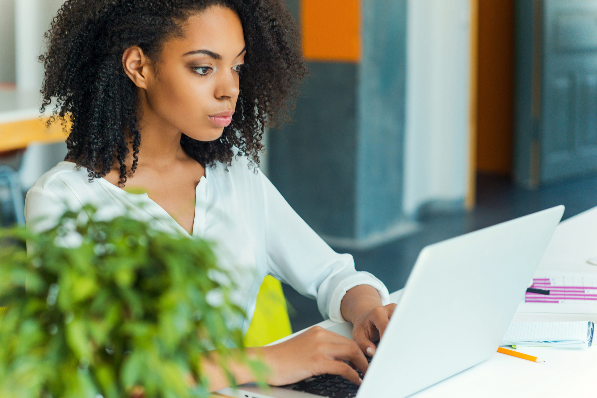 A young woman with curly hair sits concentrating on a laptop computer. She wears a white blouse and sits in a modern office environment. In the foreground, a green plant is vaguely visible.