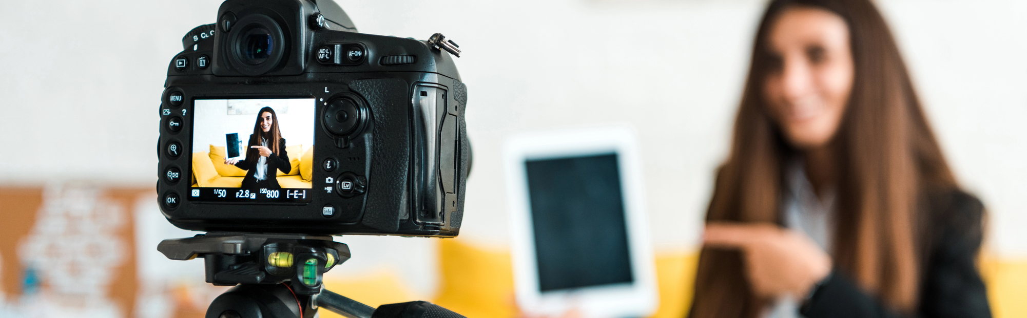 "A woman with long brown hair presents a tablet while standing in front of a camera. The camera is focused on the screen, on which the woman is visible while pointing at the tablet. The background is out of focus and contains yellow elements."