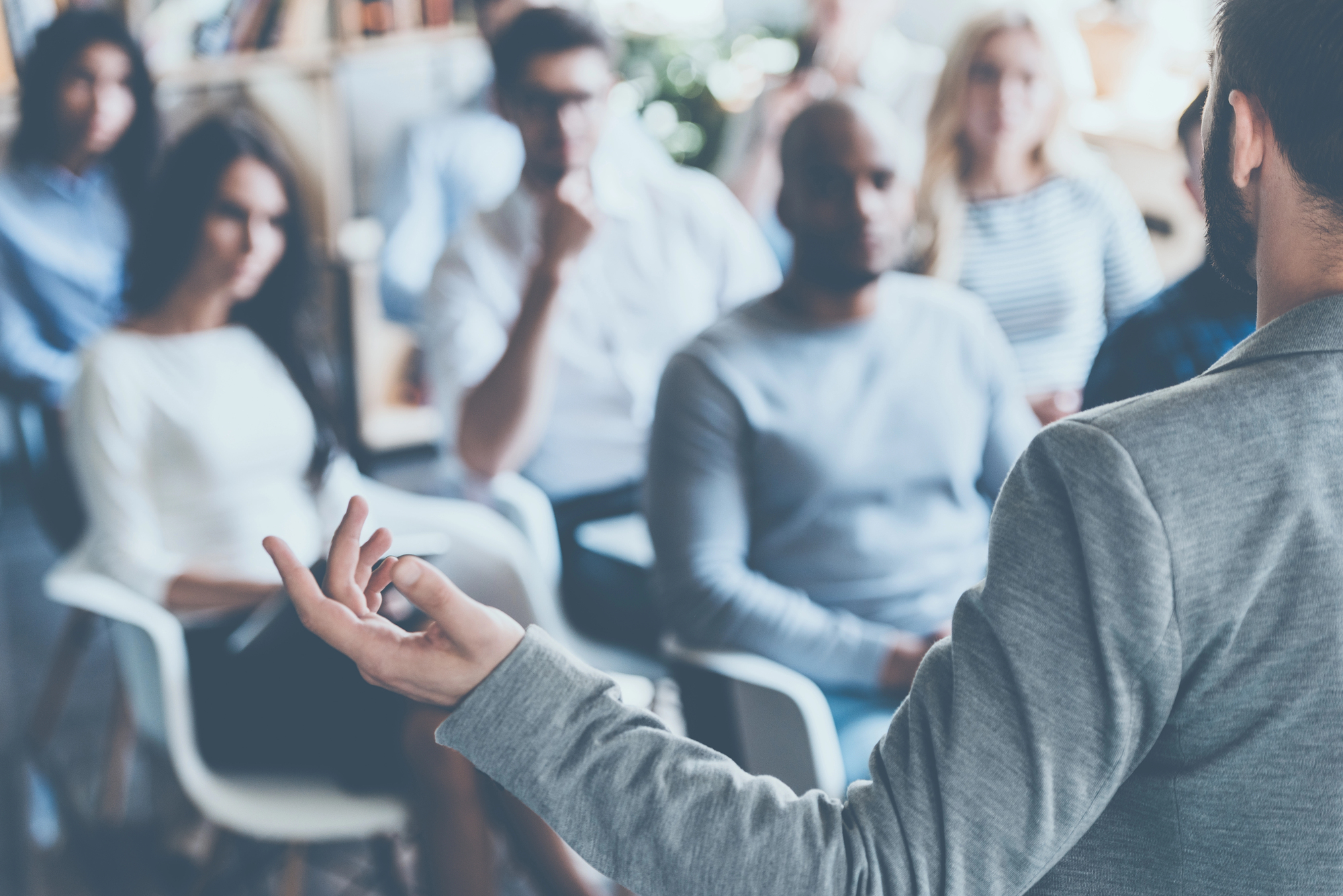 A group of people listen intently to a speaker during a presentation or workshop. The speaker, seen from the back, gestures with his hand as he speaks. The listeners, seated in modern chairs, watch the speaker with focus.