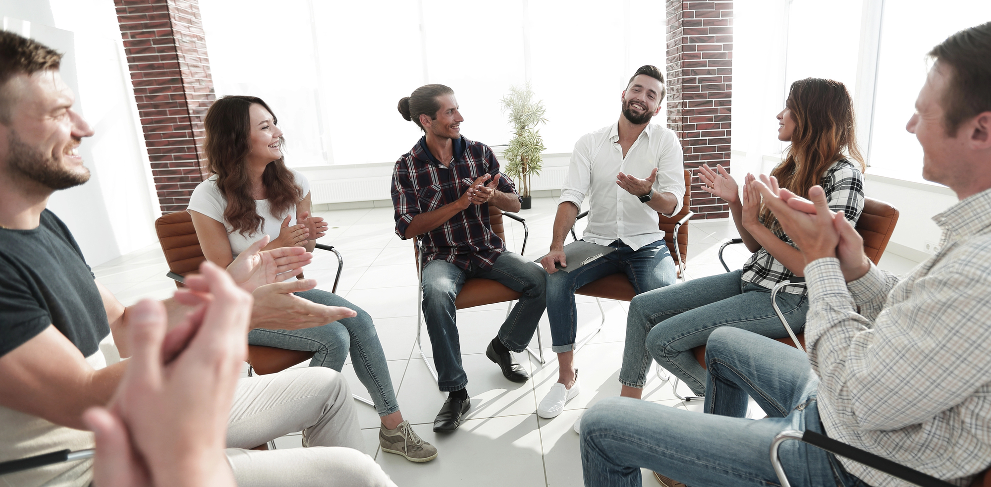 A group of people sit in a circle. They laugh and applaud. The room is bright and modern, with large windows and brick walls. Everyone sits on chairs in an open arrangement.