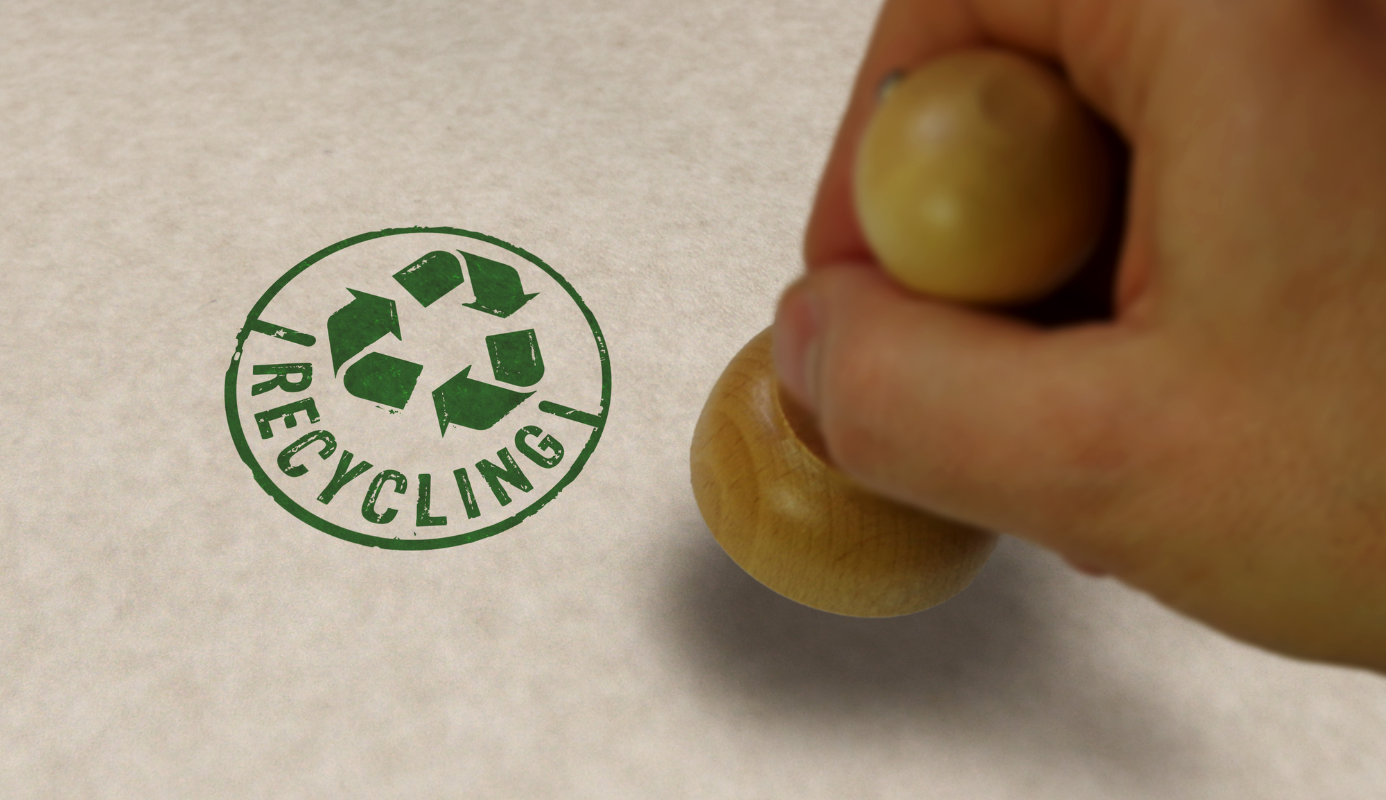 A photograph of a hand pressing a stamp on paper. The stamp contains a green recycling symbol and the word "RECYCLING" underneath.