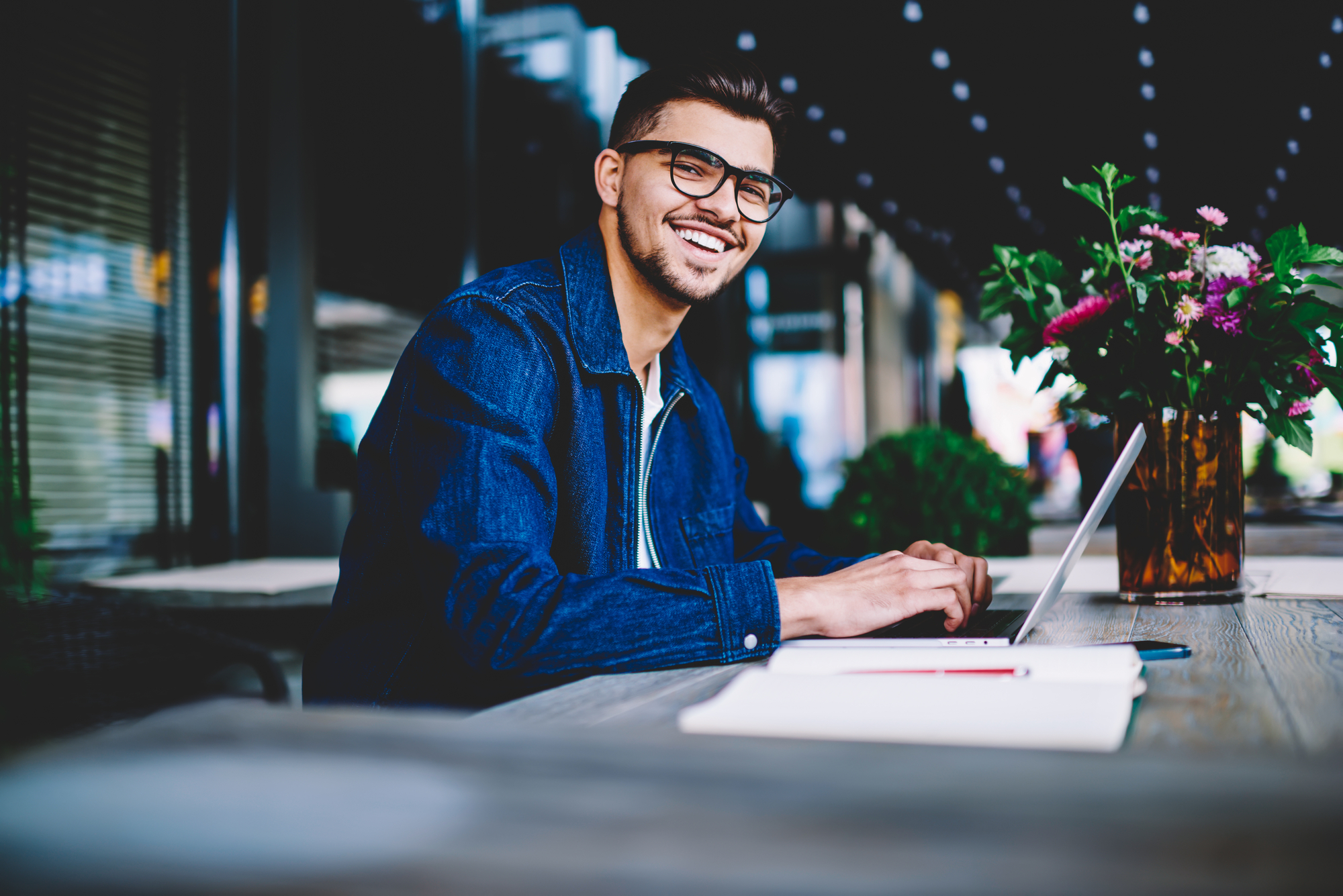 a man wearing a blue blouse and glasses sits behind a laptop and smiles into the camera