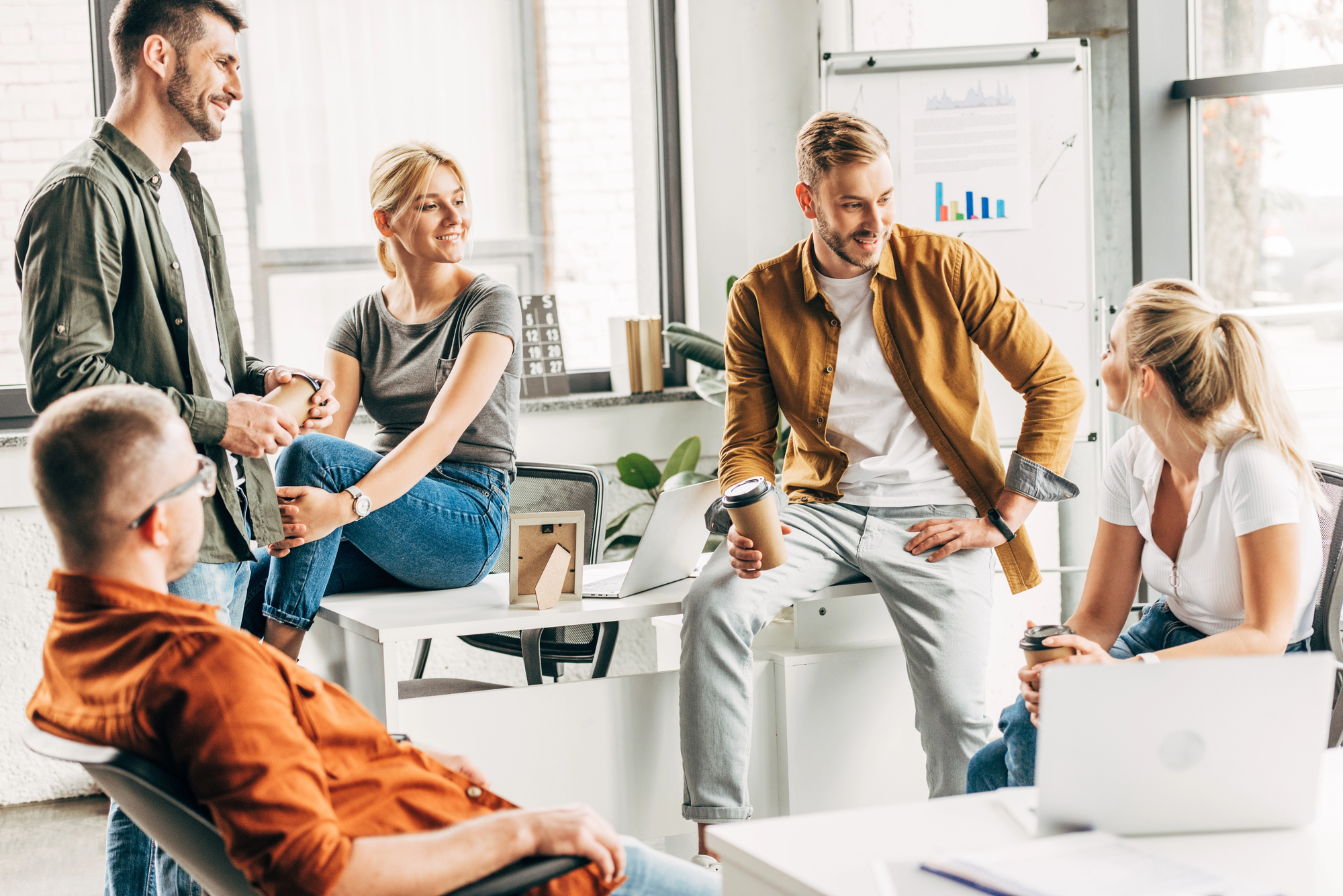 Group of young professionals in casual attire meeting informally in a bright office space, with a whiteboard in the background displaying charts. They appear to be involved in a relaxed discussion or brainstorming session.