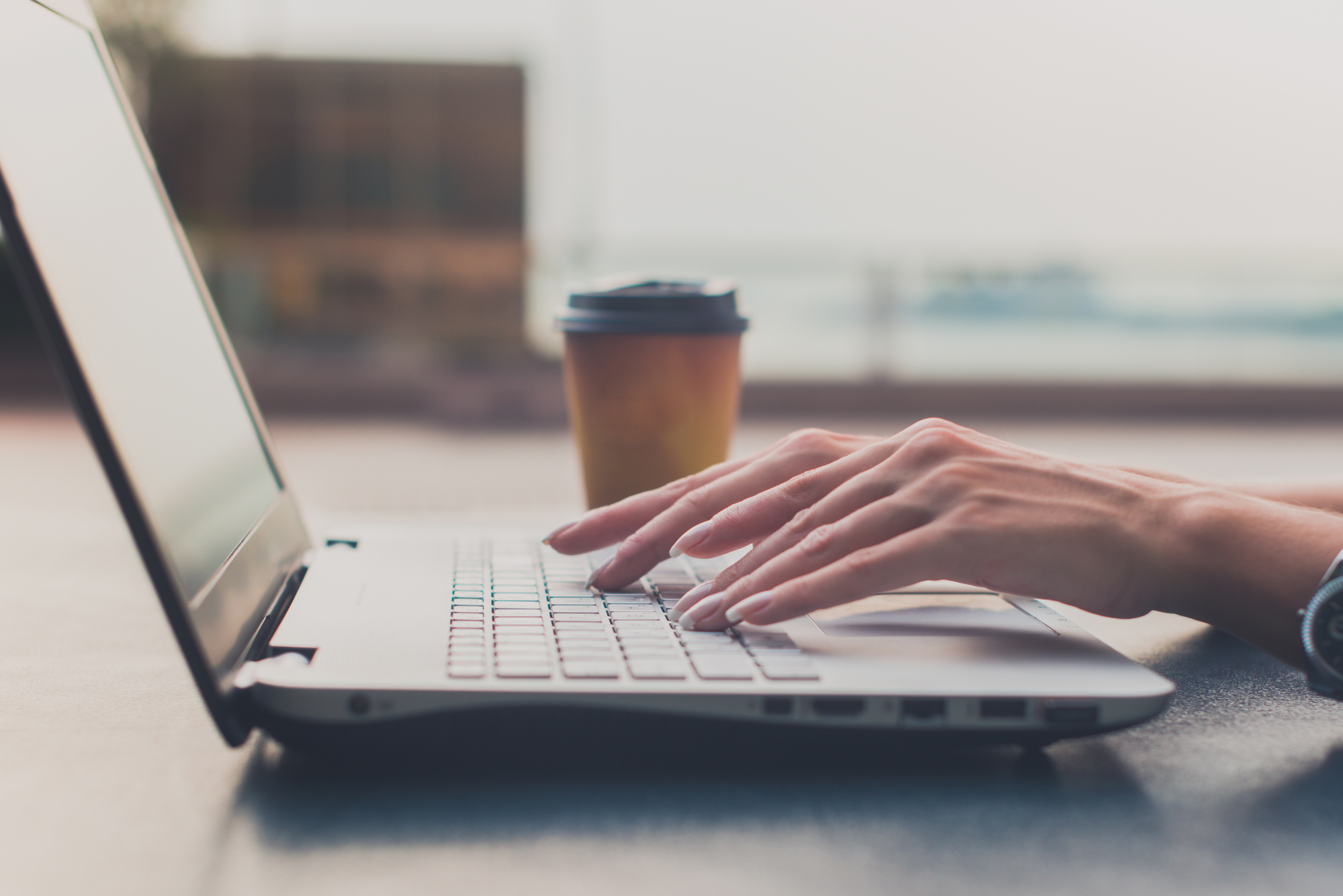 The hands of a person typing on a laptop keyboard. Next to the laptop is a coffee cup with a lid. The background is blurred but shows a calm sea and sky, suggesting that the person is working outdoors at a table, possibly on a terrace or balcony overlooking the water. This image creates an atmosphere of relaxed work in an inspiring environment.