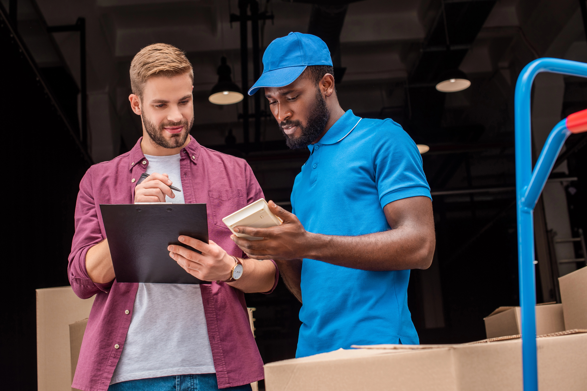 Two men in a warehouse or storage area are looking at papers together. One man in a purple shirt holds a clipboard, while the other man in a blue uniform with cap holds a notebook. Boxes and a blue trolley are visible in the background.