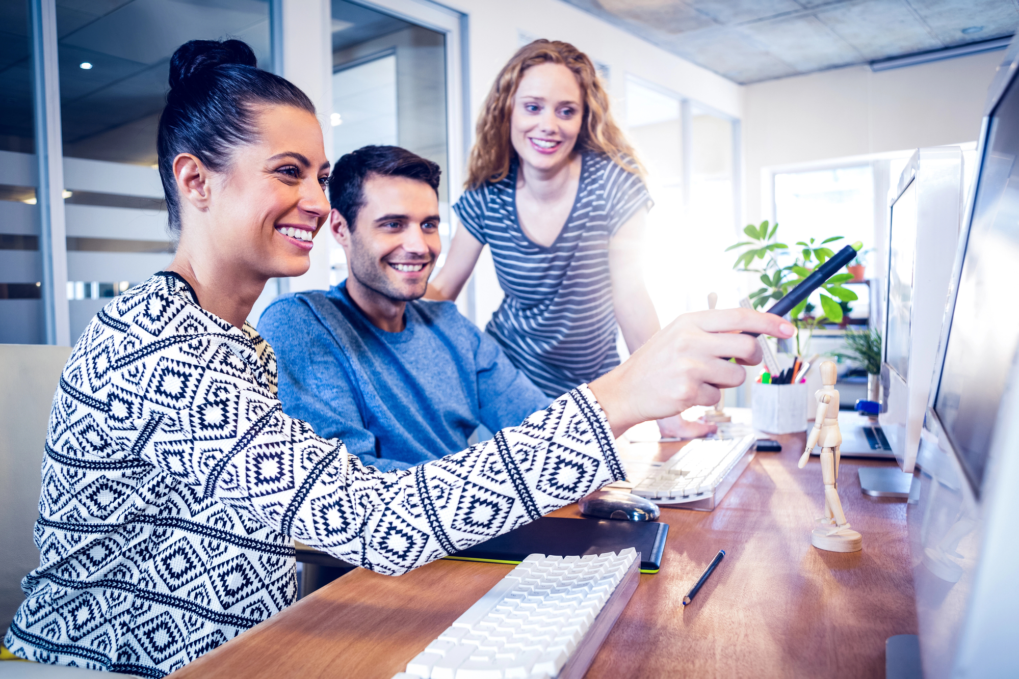 Three people are working together on a project in an office. A woman with a bun and a black and white patterned sweater points to a computer screen with a pen, while a man next to her and a woman behind them look on and smile. On the desk are keyboards, a mouse, pens and a wooden model doll.