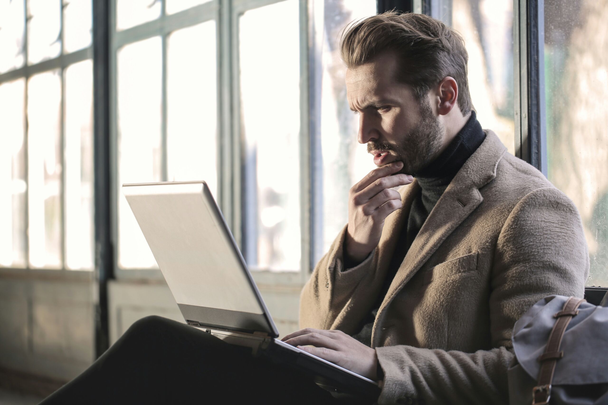 A man in a beige coat sits by a window and looks at his laptop with a pensive expression. He has his hand against his chin, suggesting he is deep in thought or trying to solve a problem. The natural light from the window falls on him, creating a contemplative atmosphere. His clothing and posture give a professional impression.