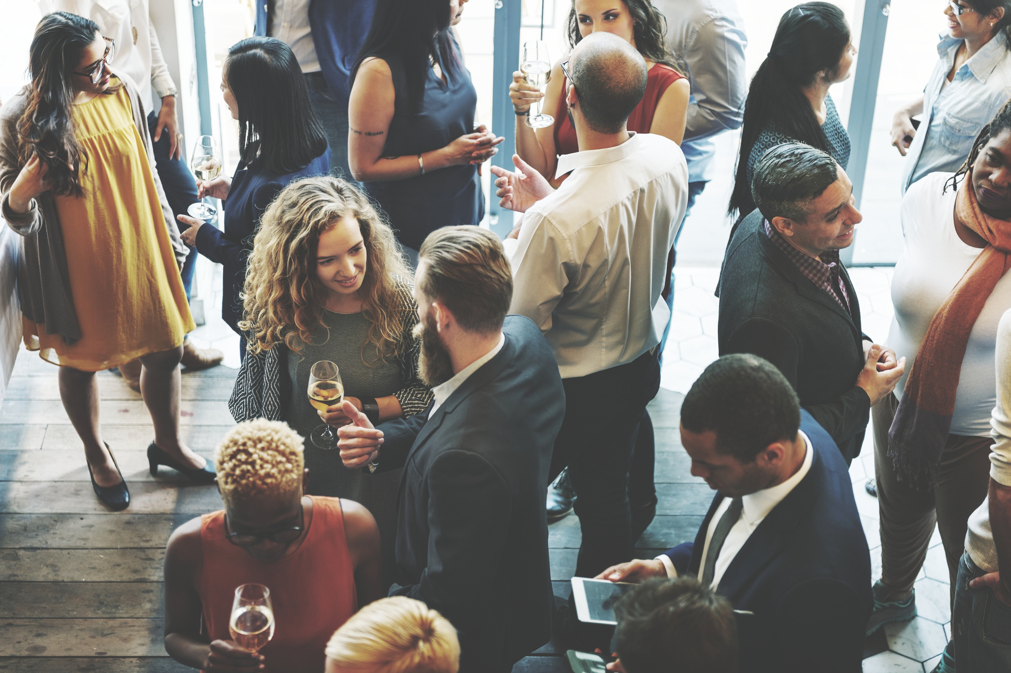 Diverse group of professionals in business attire socializes during a networking reception, wine glasses in hand in a room with large windows and wooden floor.