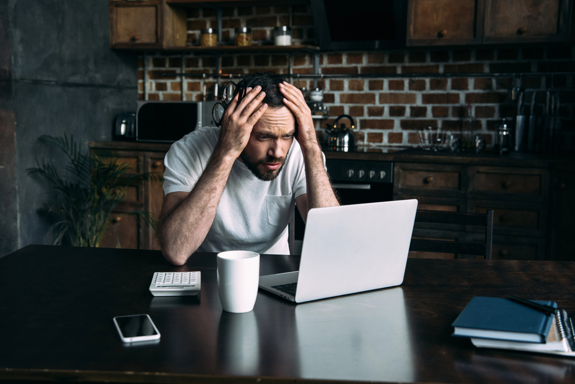 A man in a white t-shirt sits at a table in a kitchen, hands on his head as he looks at a laptop with a frustrated expression. Also on the table are a coffee cup, calculator, smartphone and notebook.