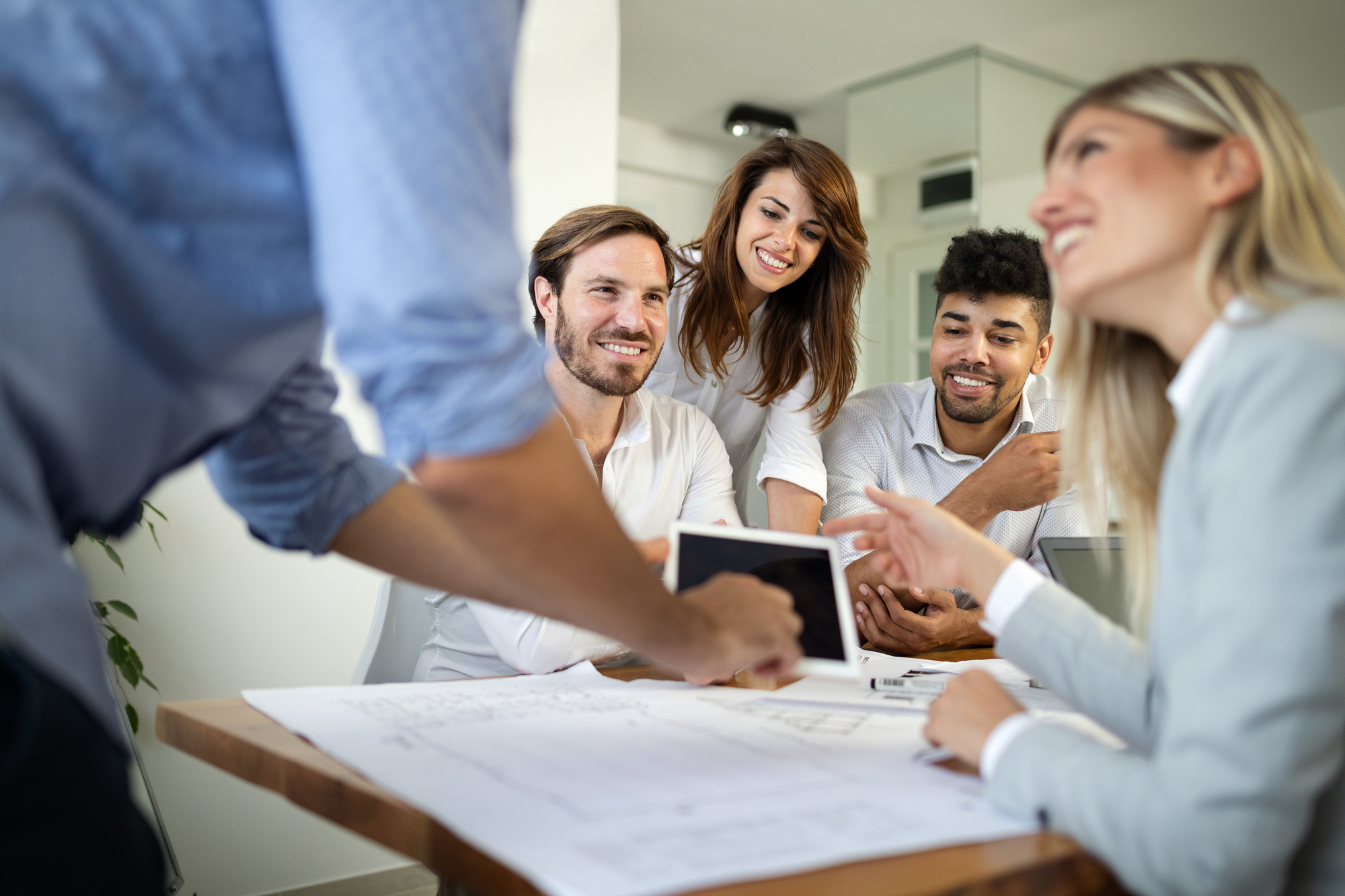 A diverse group of smiling young professionals in a revenue team meeting reviews documents and tablets together at a conference table.