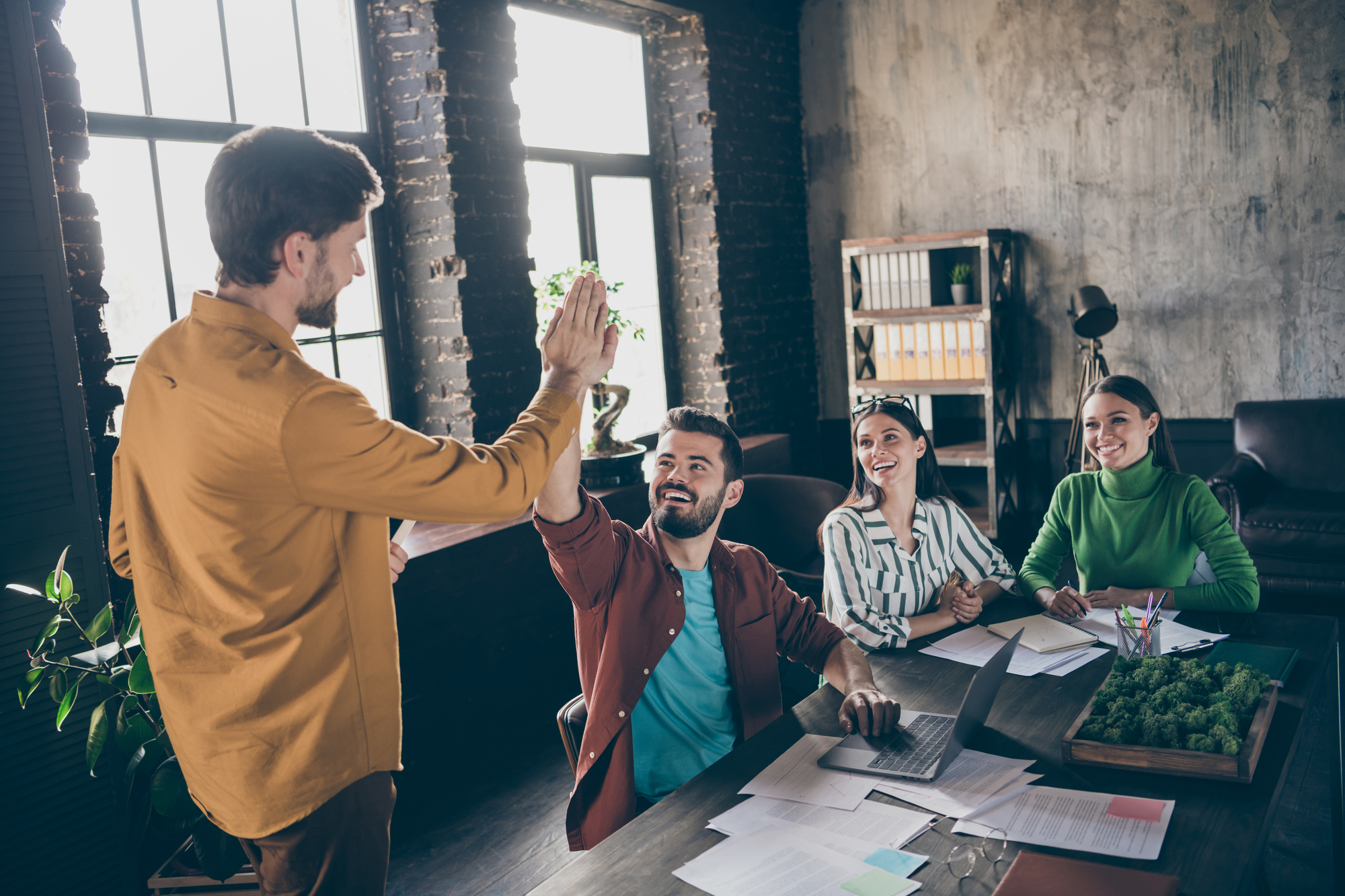 Four people in a modern office. A man gives a high-five to another man, while two women look on smiling. The office has an industrial style with large windows and brick walls.