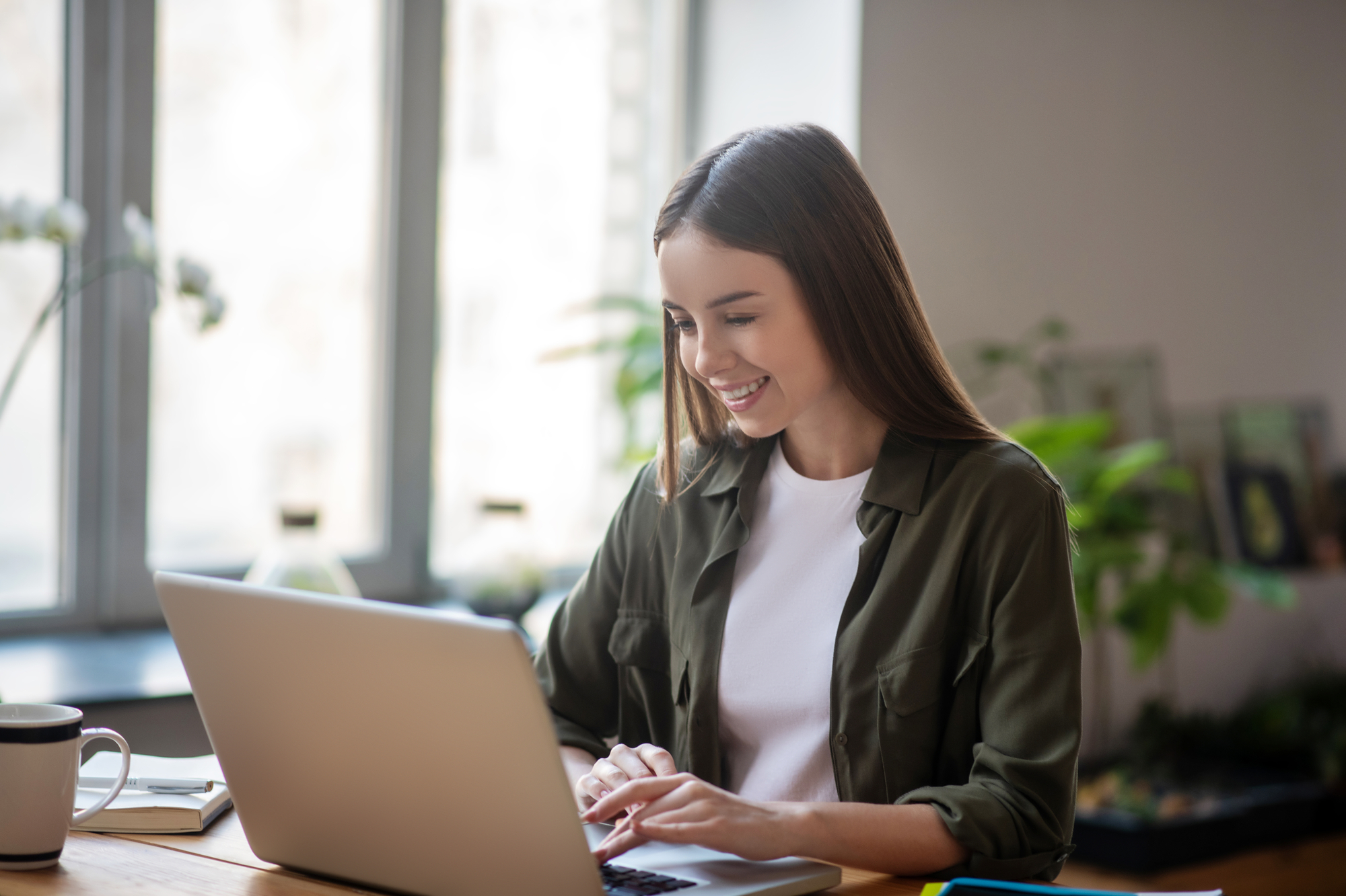 Image of a young woman smiling as she works on her laptop. She is sitting at a wooden table with a cup of coffee and notebook beside her. A window and some houseplants can be seen in the background.