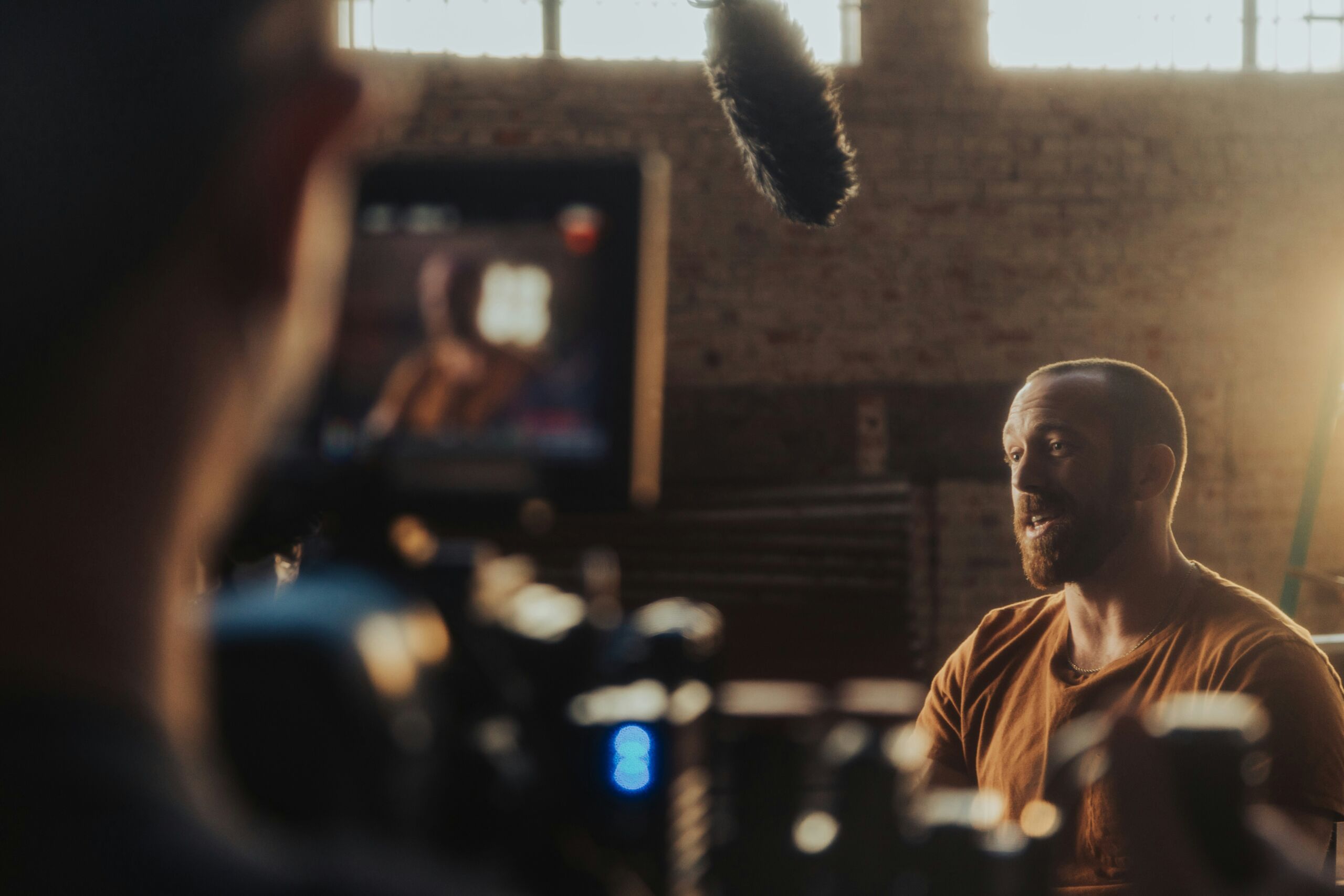 Man with beard in orange shirt is interviewed on camera in studio with natural light and visible microphone boom