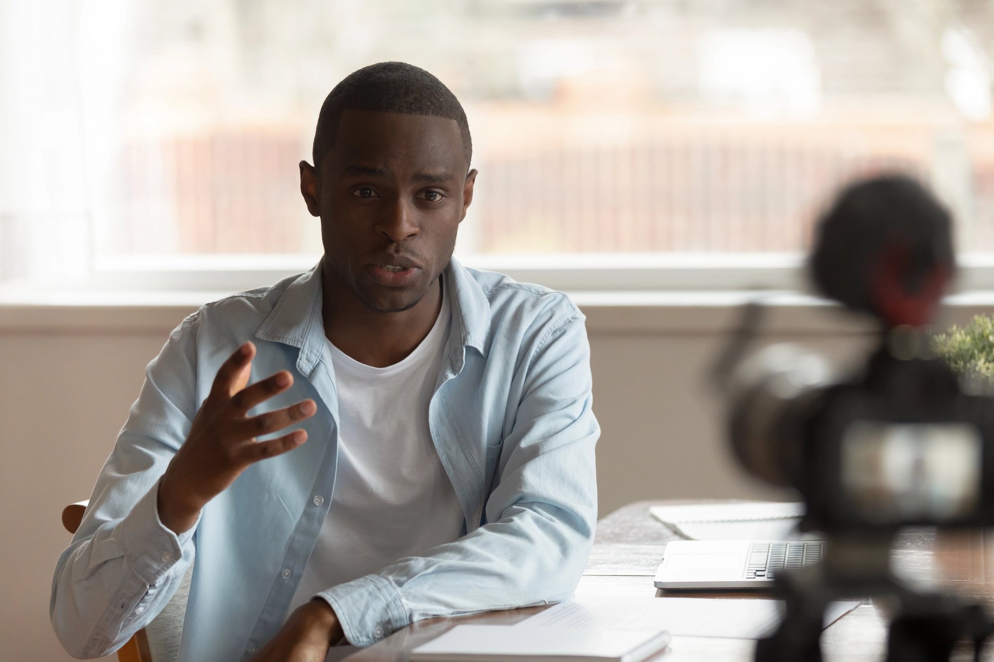 A man in a light blue shirt and white t-shirt sits at a desk and speaks toward a camera, using an expressive hand movement.