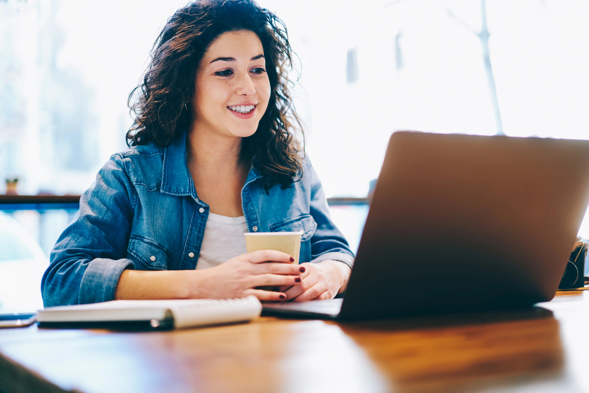 dark-haired woman with denim blouse sits behind a laptop with a cup