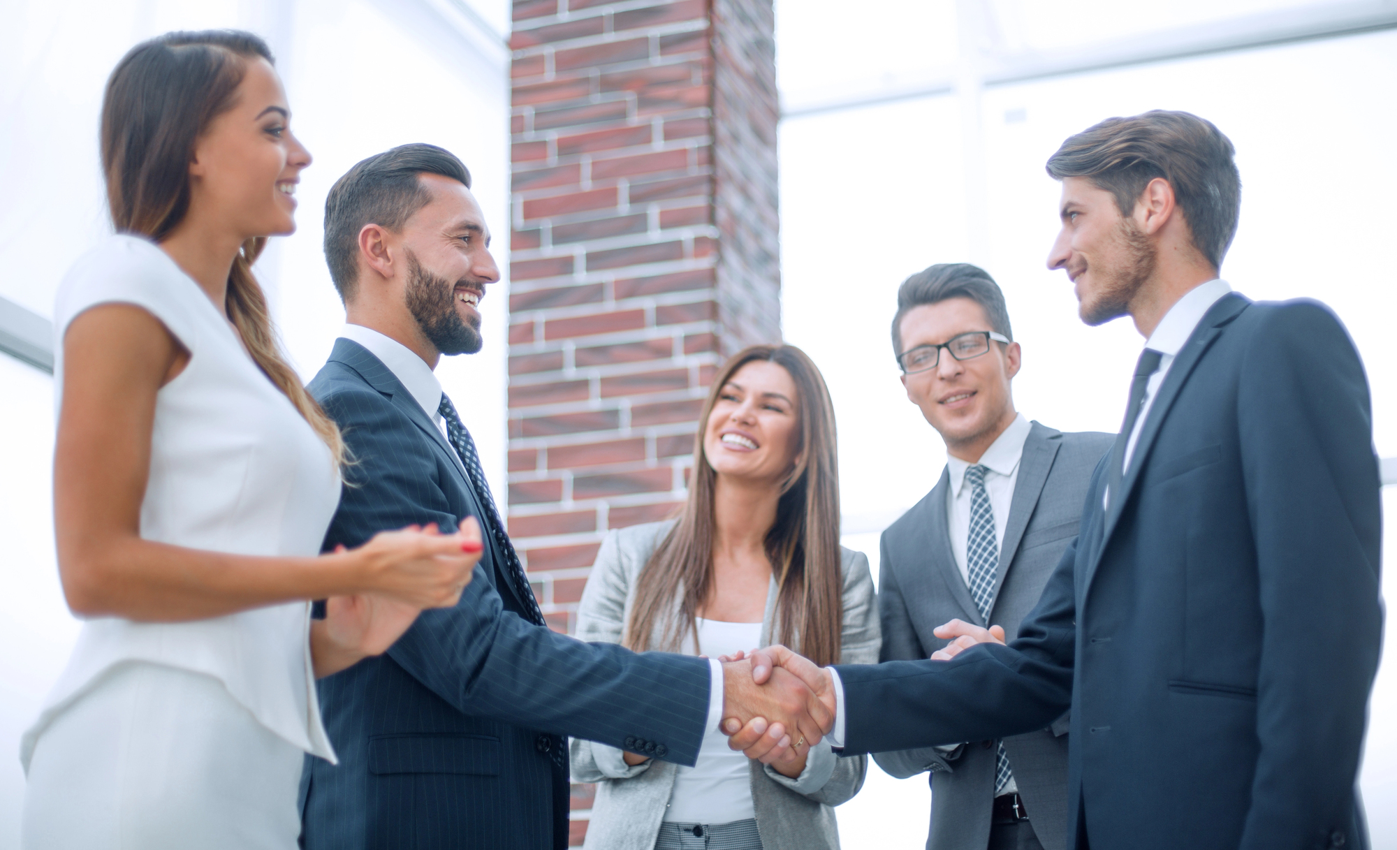 A group of five young professionals in business attire stand in an office setting with a brick wall in the background. Two men shake hands while three other colleagues stand there smiling. One woman wears a white dress, another wears a gray jacket, and the men wear dark suits with ties. One of the men wears glasses.