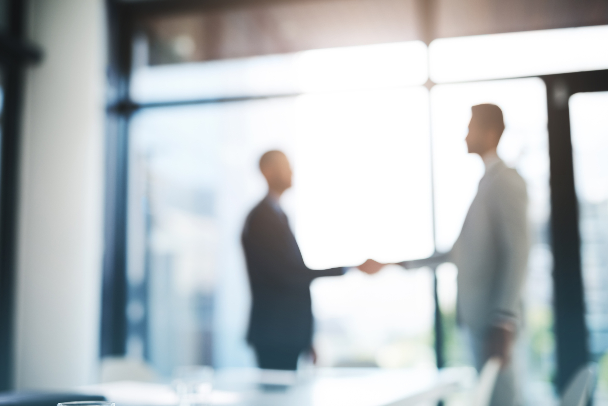 Image of two businessmen shaking hands in a modern office setting. The photo is out of focus, blurring the details of the people and their surroundings, but the handshake remains clearly visible.