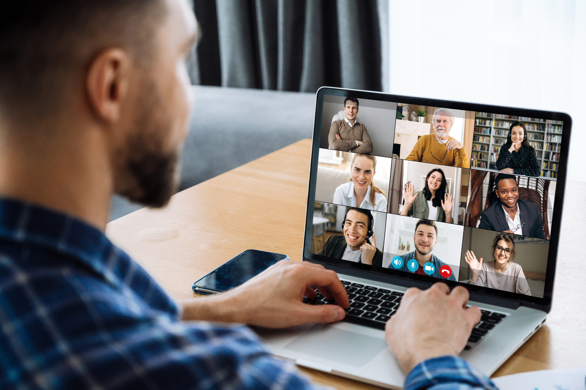 Man participates in a video call on his laptop. On the screen, nine other participants are visible smiling and waving at the camera. The participants vary in age and background. The man is wearing a plaid blouse and has a phone next to his laptop.