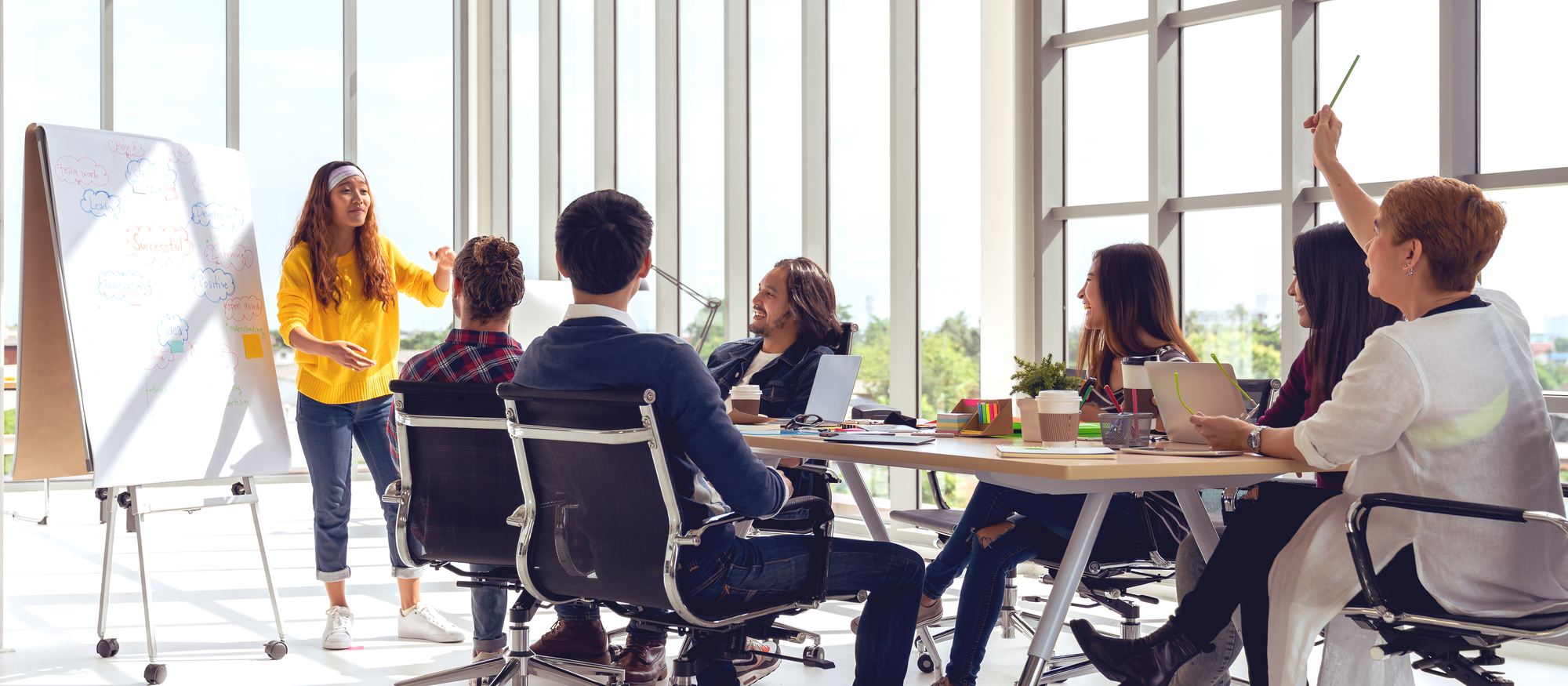 A group of six people is in a bright, modern office space, gathered around a table for a meeting or workshop. A woman in a yellow sweater stands at a whiteboard, presenting and engaging the group. The whiteboard is full of colorful notes and diagrams. The seated participants, both men and women, seem attentive and engaged, some smiling and taking notes, while one woman raises her hand to ask a question. Large windows in the background let in lots of natural light.