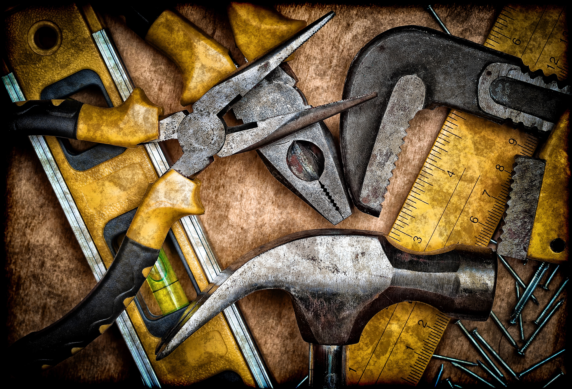 A collection of hand tools on a wooden background. The tools include a level, a pair of pliers, an adjustable wrench, a hammer, a saw and a ruler. The handles of some of the tools are yellow with black, while the metal parts show signs of use and wear. A few nails are scattered around the tools.