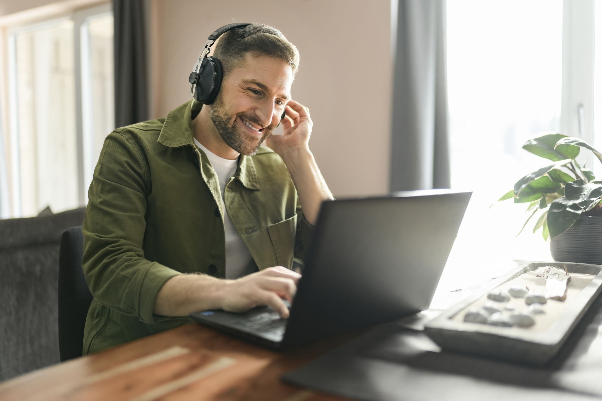 Man in green shirt sits behind laptop and smiles. He wears a headset with microphone and sits in a bright room.