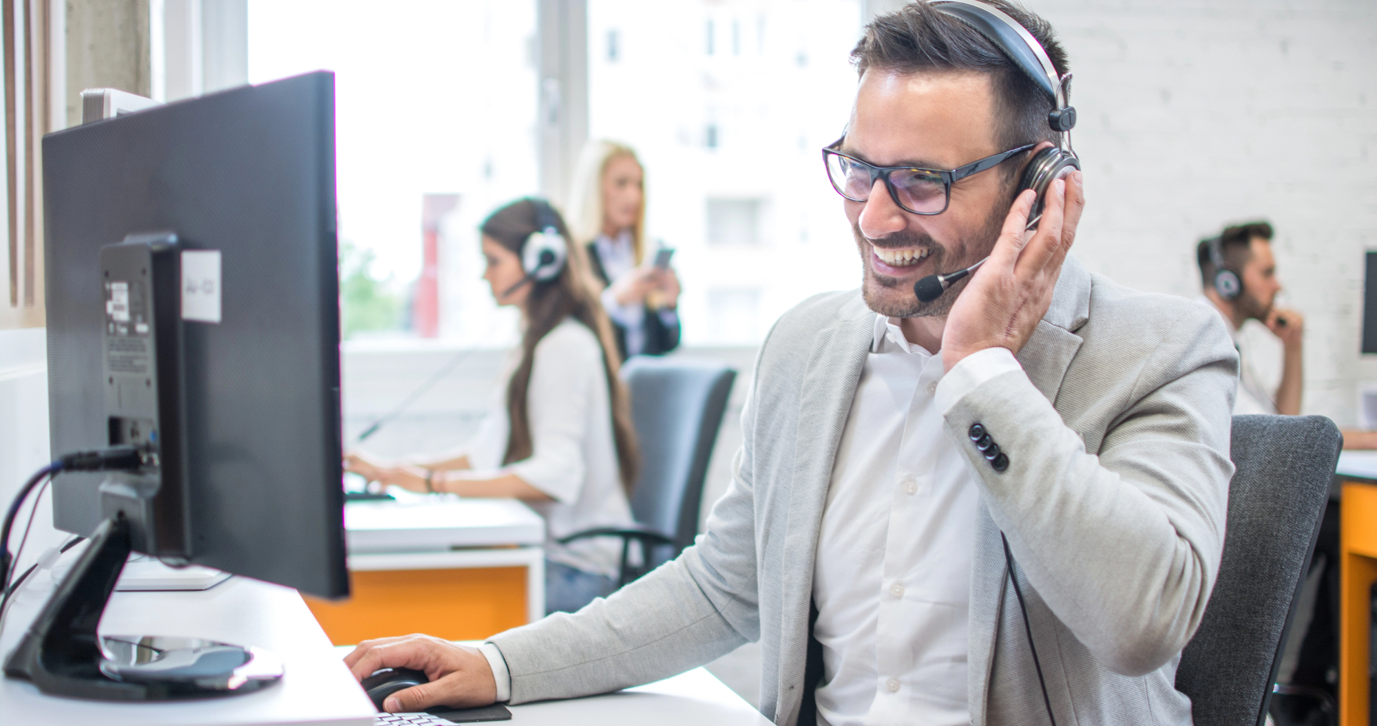 Smiling sales professional in gray jacket and headset behind computer screen in open office space, with colleagues in background.