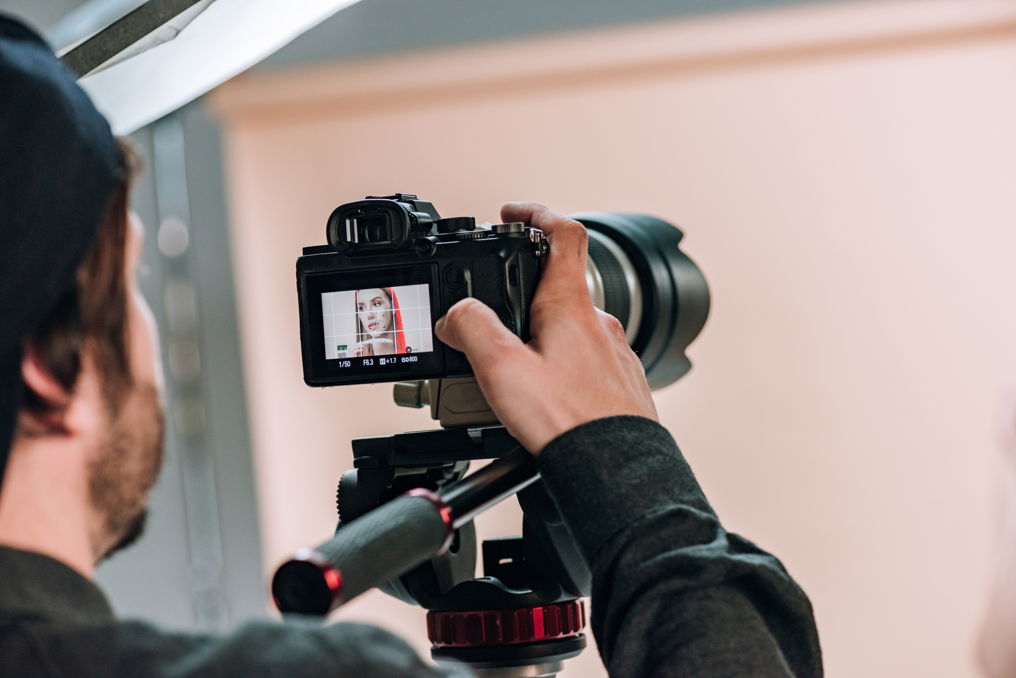 Close-up of a cameraman shooting video. The camera's display shows a woman wearing a red scarf, brought into sharp focus. The cameraman is wearing a dark garment and a cap while holding the camera and adjusting the focus. The background is neutral and light in color.