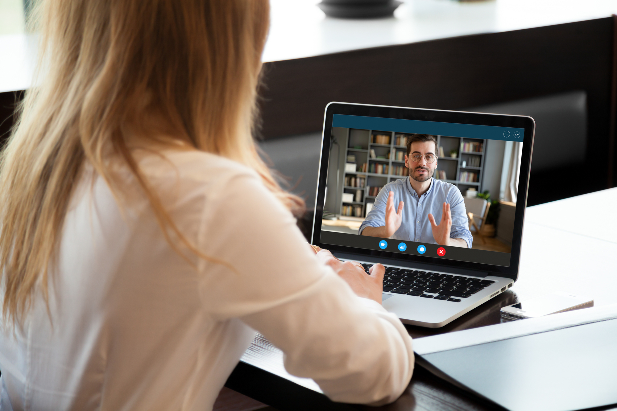 A woman in a white shirt watches a video call on her laptop, in which a man with glasses and beard in a light sweater sits in front of a bookcase talking and making hand gestures.