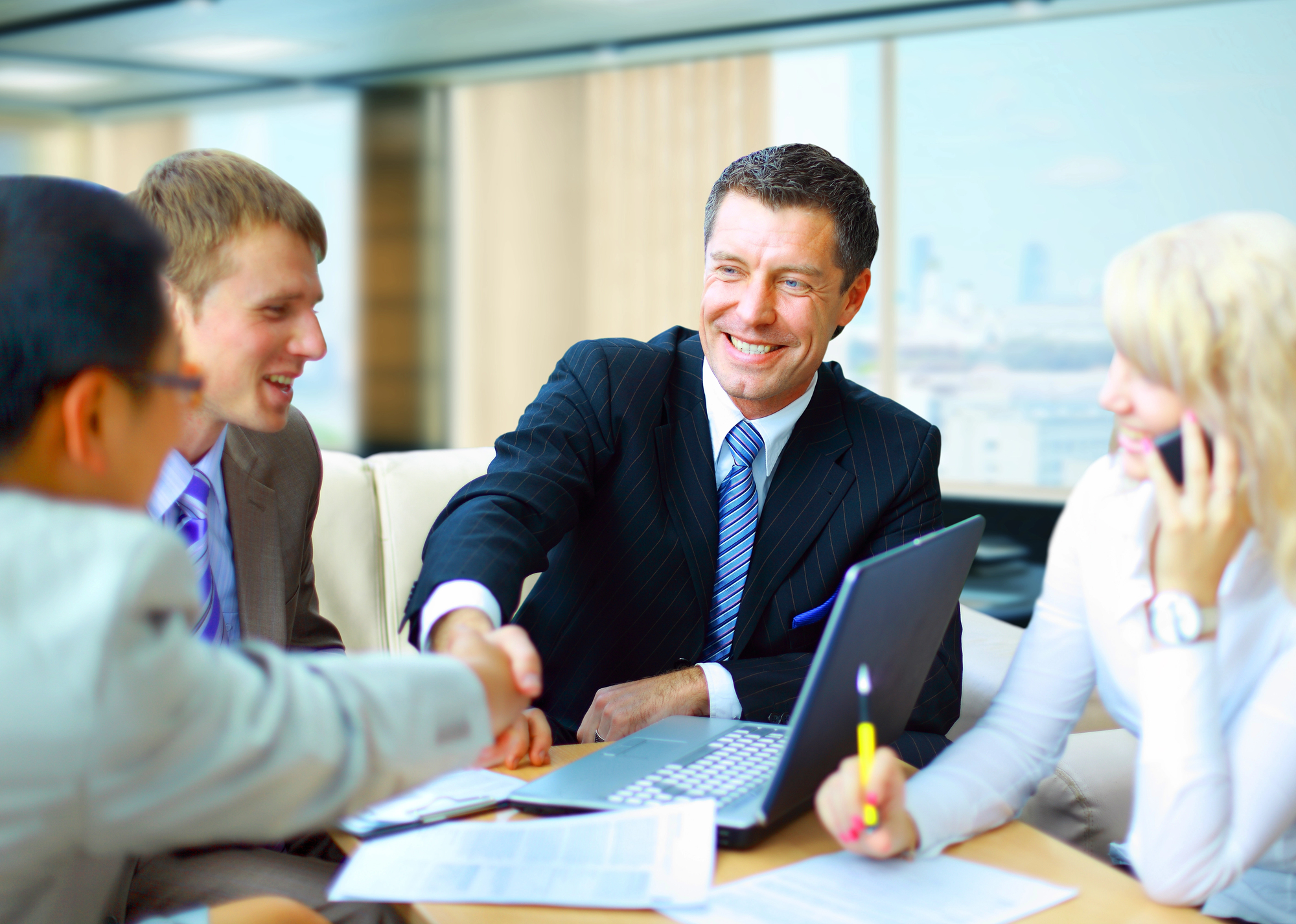 A group of businessmen holding a meeting in a modern office setting. A man in a dark suit and tie smiles and shakes another person's hand, while documents and a laptop are on the table in front of them. A woman on the right talks on the phone and holds a pen. The atmosphere looks professional and friendly, indicating a successful business partnership or deal.