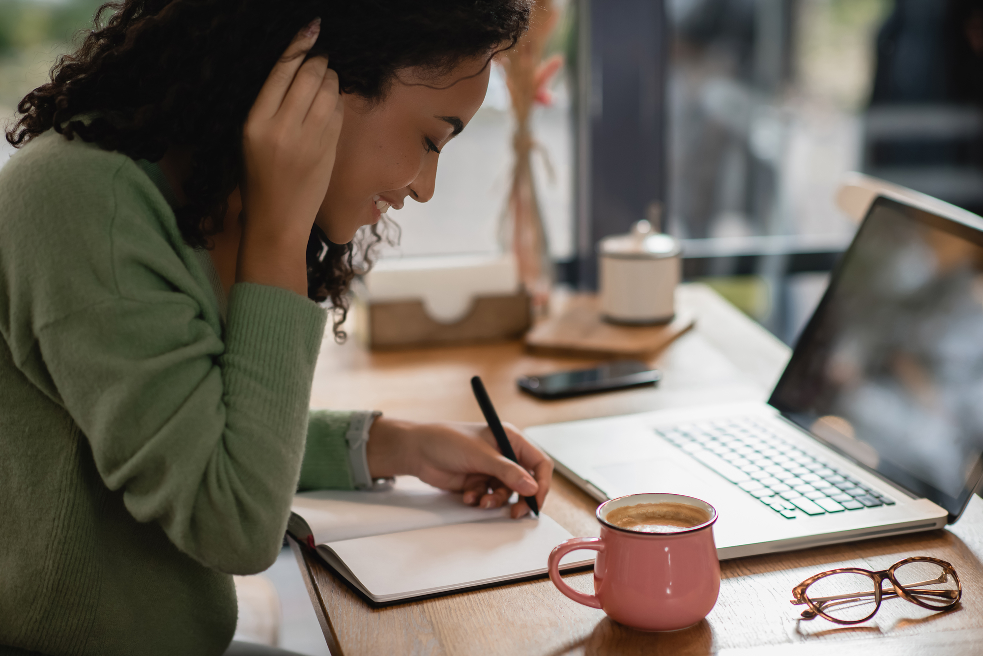 A young woman with curly hair sits at a wooden table in a café. She wears a green sweater and writes in a notebook with a pen. In front of her are a laptop and a pink coffee cup. Her glasses and smartphone are also on the table.