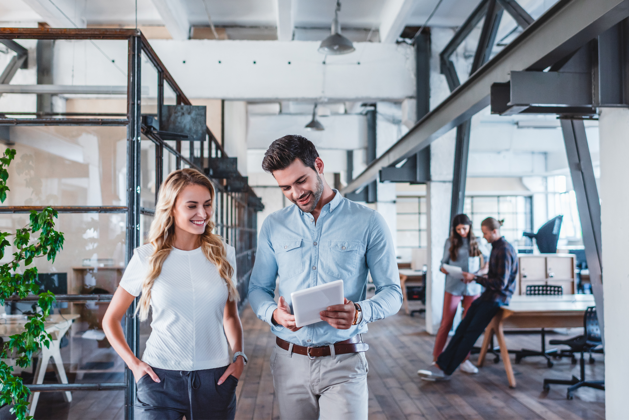 a woman wearing a white blouse and a man wearing a light blue shirt are standing in an open office space. The man has a tablet in his hand.